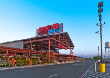 Exterior of an H-E-B Plus grocery store at dusk, with the illuminated H-E-B Plus sign, a garden center displaying potted plants and flowers along the front entrance, and an empty parking lot in the foreground