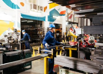 Two workers in blue coveralls and hard hats cutting steel structural sections in a metal fabrication facility