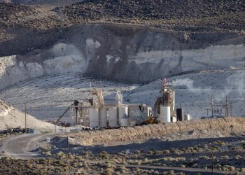 Industrial rare earth mineral processing facility with silos and processing towers set against white desert cliffs, American flag flying above the complex