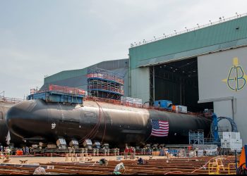 Submarine hull draped with an American flag outside General Dynamics Electric Boat's shipyard, with workers on the dry dock in the foreground