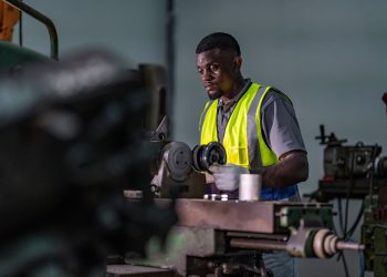 Factory worker in a safety vest inspecting metal on a lathe machine in an industrial machine shop