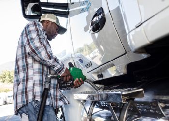 Truck driver in a plaid shirt and cap filling a white semi truck's fuel tank with a green diesel nozzle at a truck stop