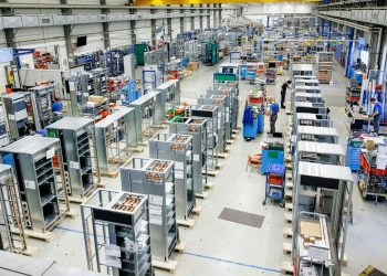 Rows of low-voltage switchgear enclosures under assembly on the production floor of Siemens' Leipzig plant, with workers at stations