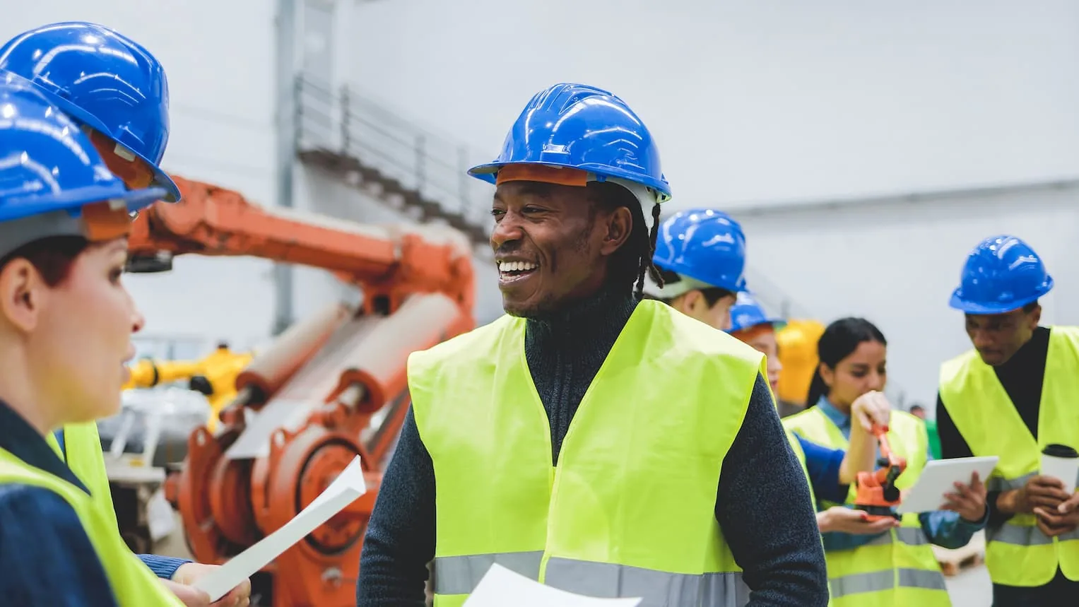 A group of workers in blue hard hats and yellow high visibility vests talking on a factory floor, with an orange industrial robot arm behind them