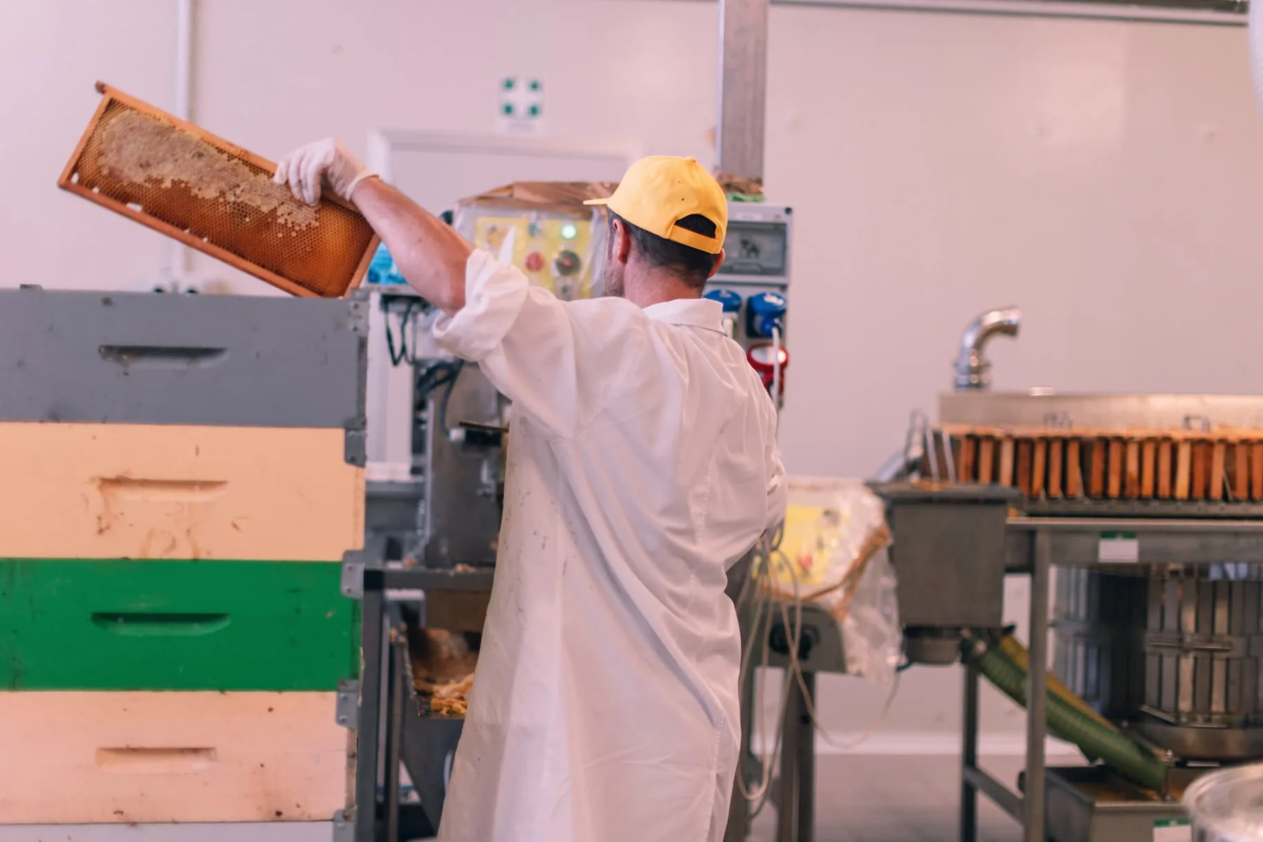 A beekeeper in a white coat lifting a honeycomb frame beside hive boxes and stainless extraction equipment at a commercial honey facility