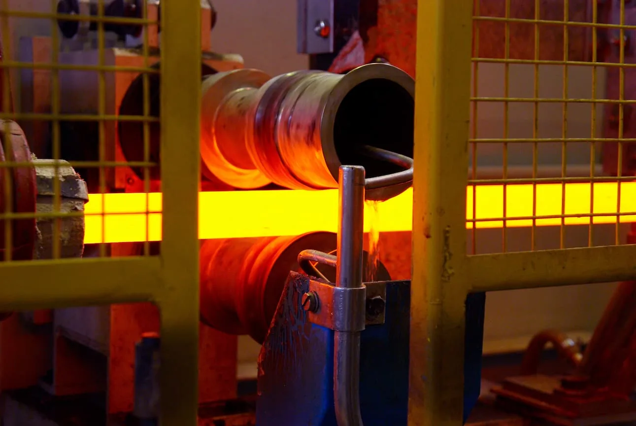 Glowing steel passing between grooved work rolls under water cooling, behind a yellow safety guard